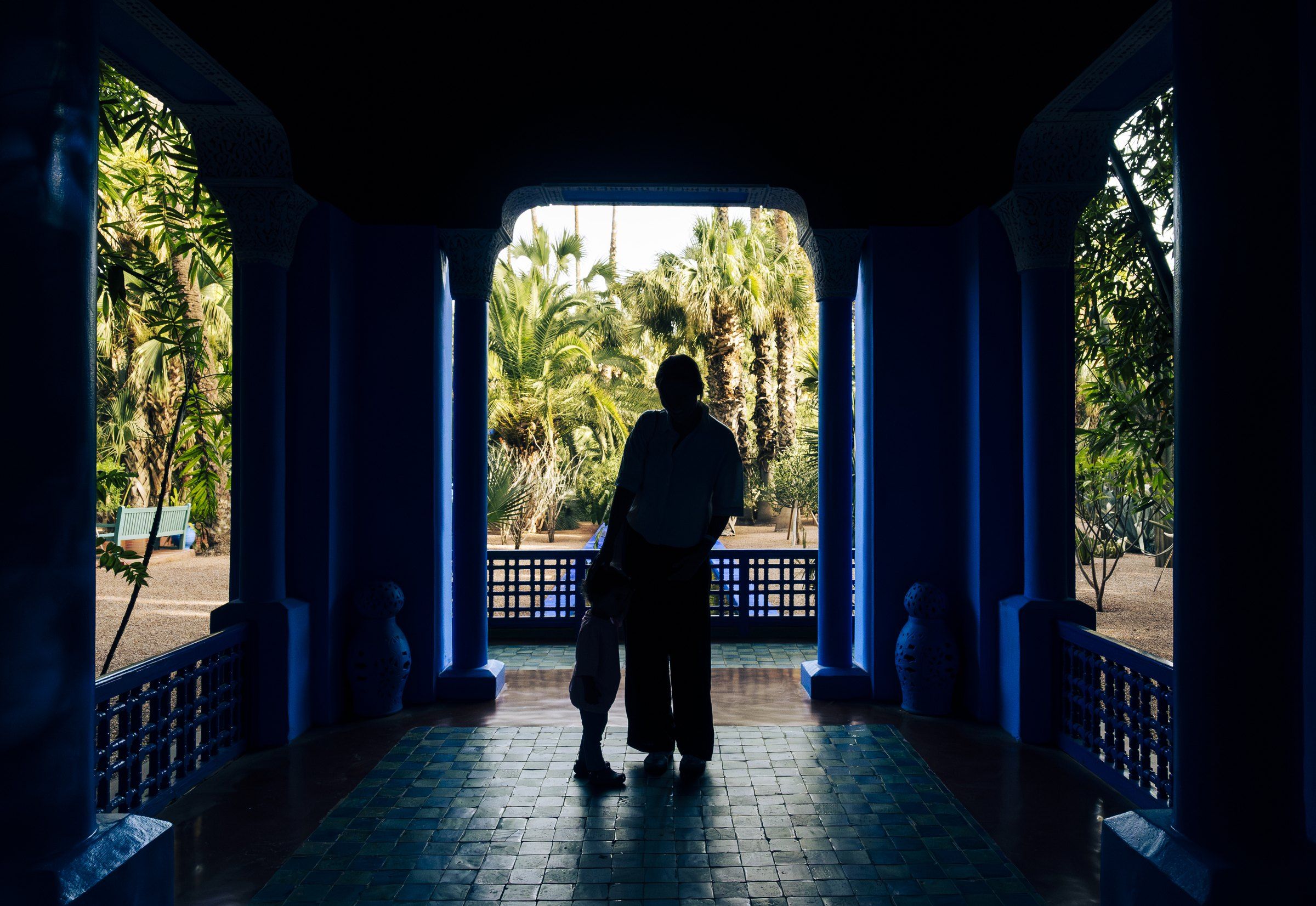 Silhouette of an adult and a small child in a cobalt-blue colonnade, framed against a sunlit garden of palms