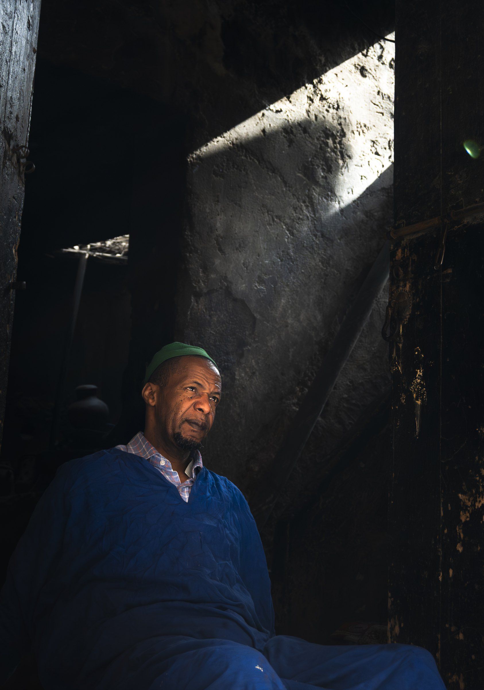 Portrait of an older man in a blue robe and green cap, lit by a shaft of light in a dark doorway