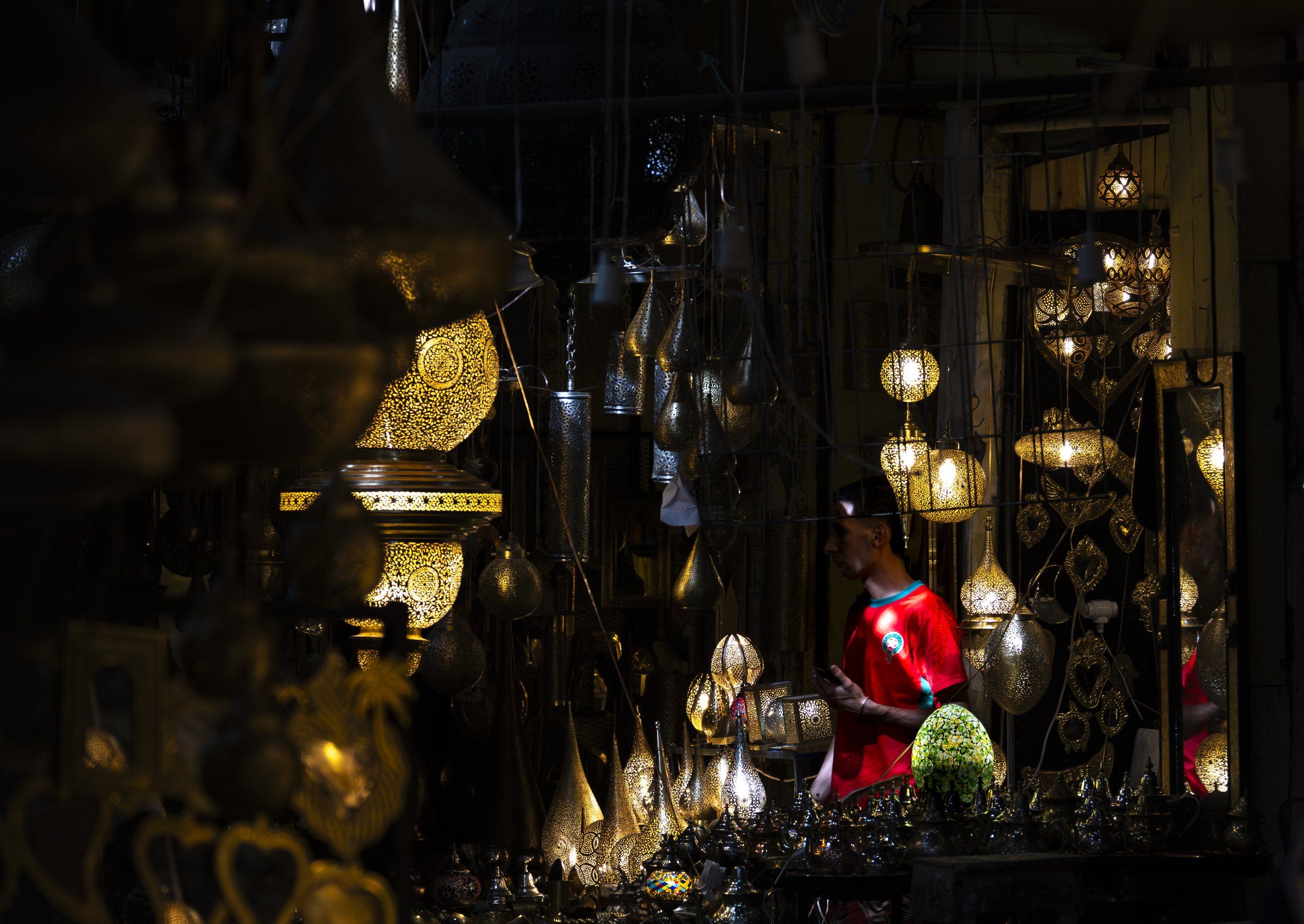 A man in a red shirt among glowing brass lanterns in a dark shop
