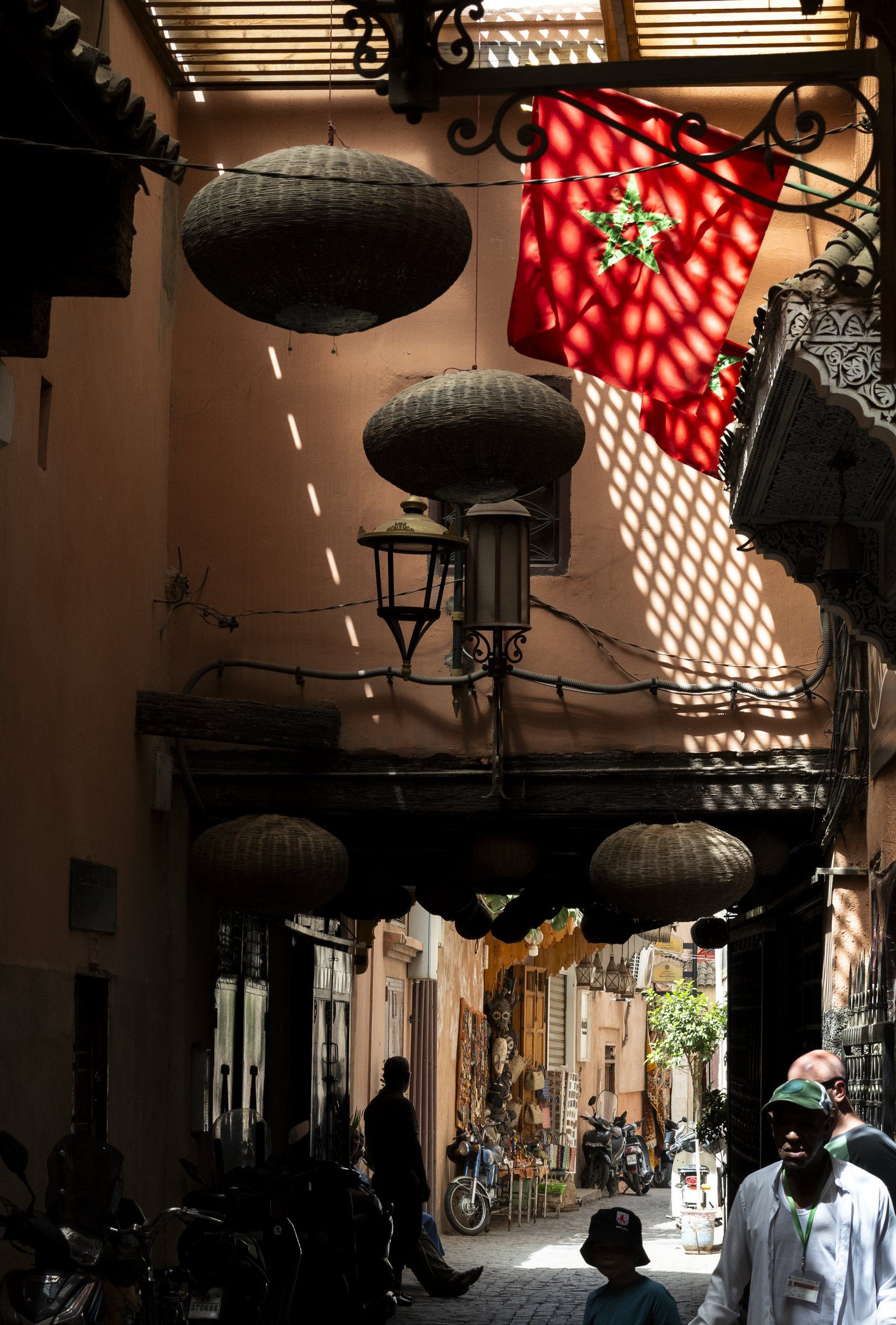 Souk street with hanging lanterns and a red Moroccan flag in dappled light, people walking below
