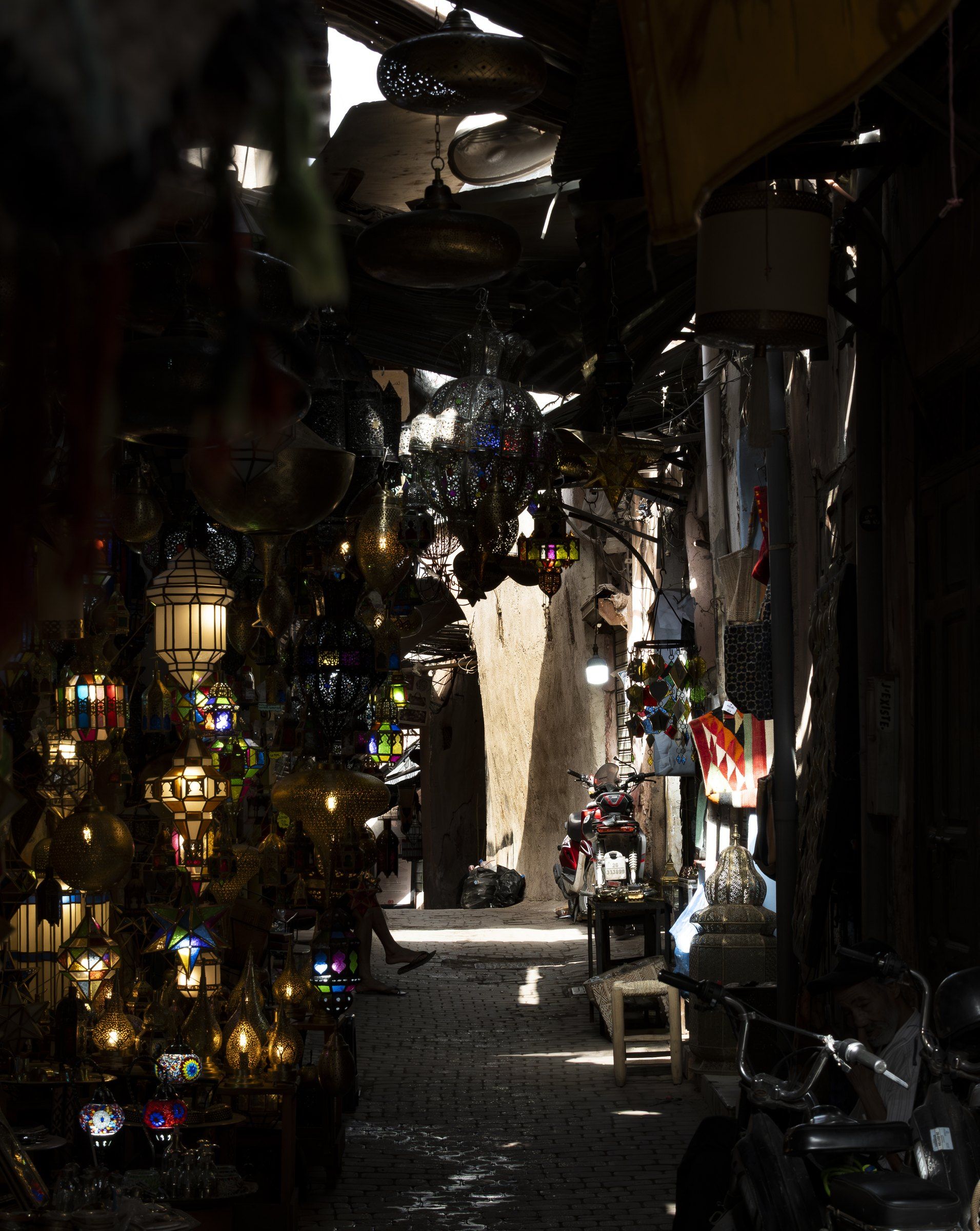 Dark souk passage lit by hanging lanterns, with daylight at the far end